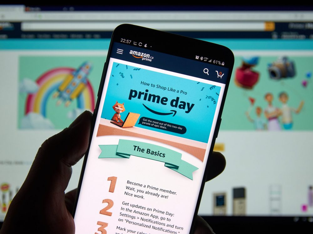 Image of a person holding a smartphone with Amazon Prime open in front of a computer screen on which Amazon is also open